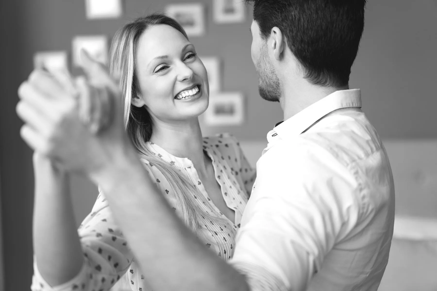 A smiling couple dances closely in a black and white scene, with a woman facing the camera and a man leading the step in a dance class setting.