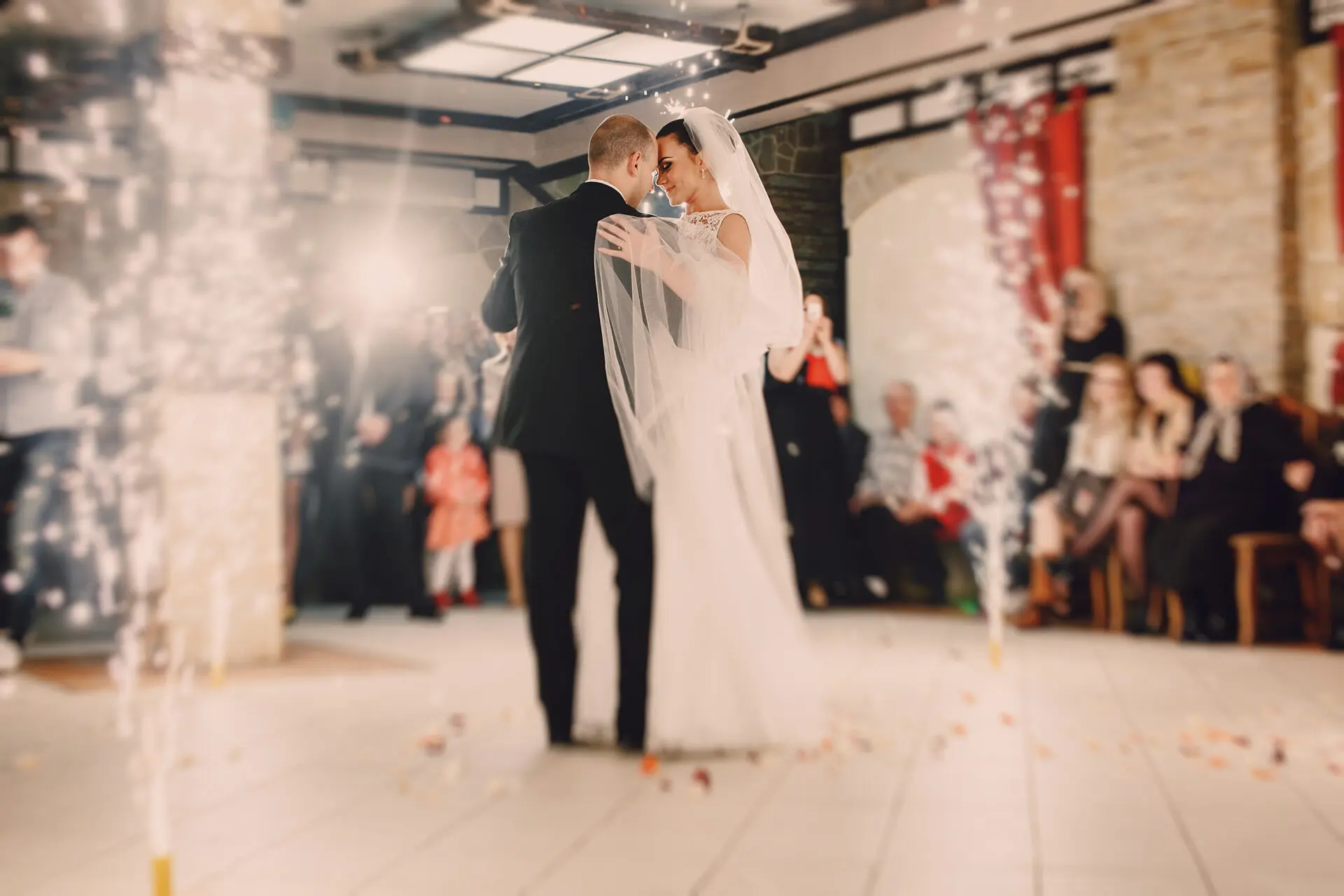 A bride and groom share their first dance in a warmly lit ballroom, with guests watching during a wedding celebration in elegant surroundings.