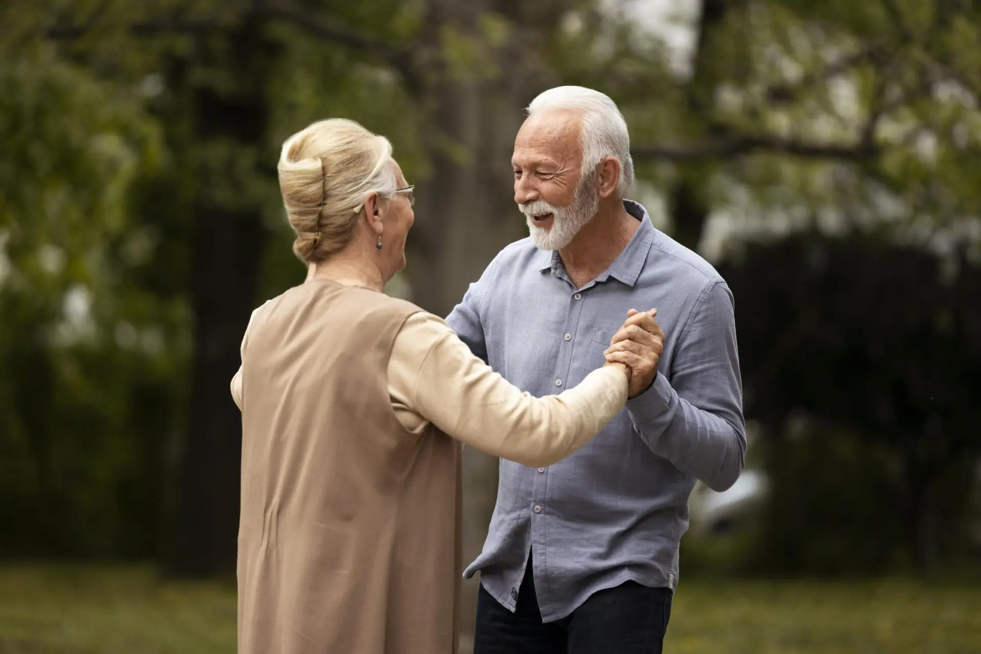 An elderly couple dances together outdoors at a warm, relaxed pace, smiling as they hold hands and move in step in the garden.