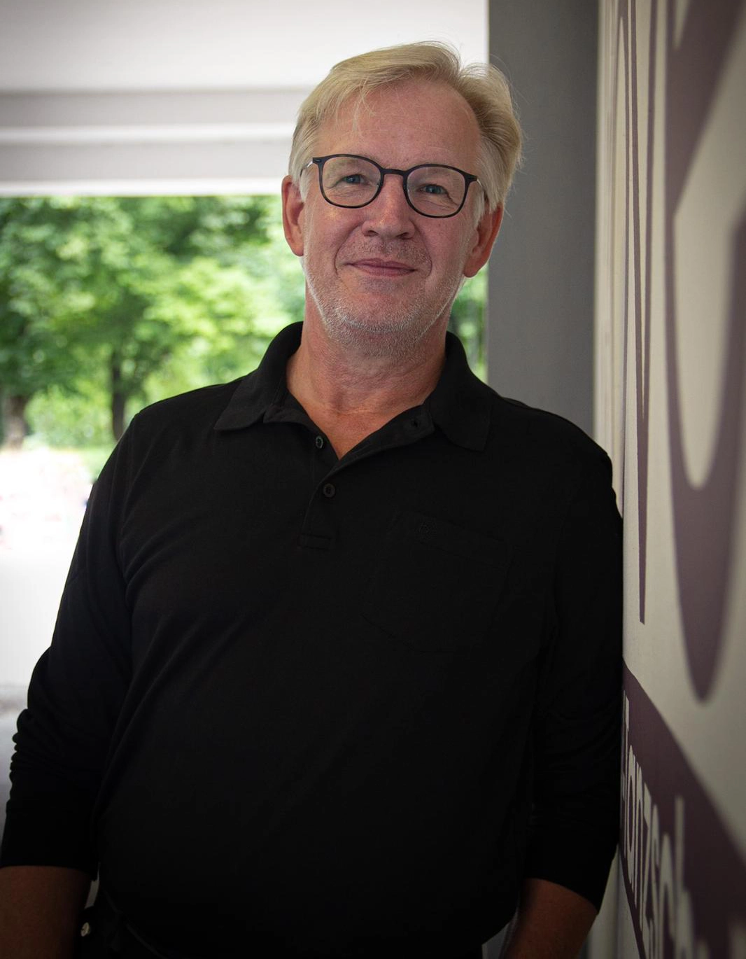 Christopher Plogmann Ein Mann namens Christopher Plogmann steht lächelnd in einem hellen Raum mit Fenster und grüner Kulisse, trägt ein schwarzes Poloshirt und Brille.