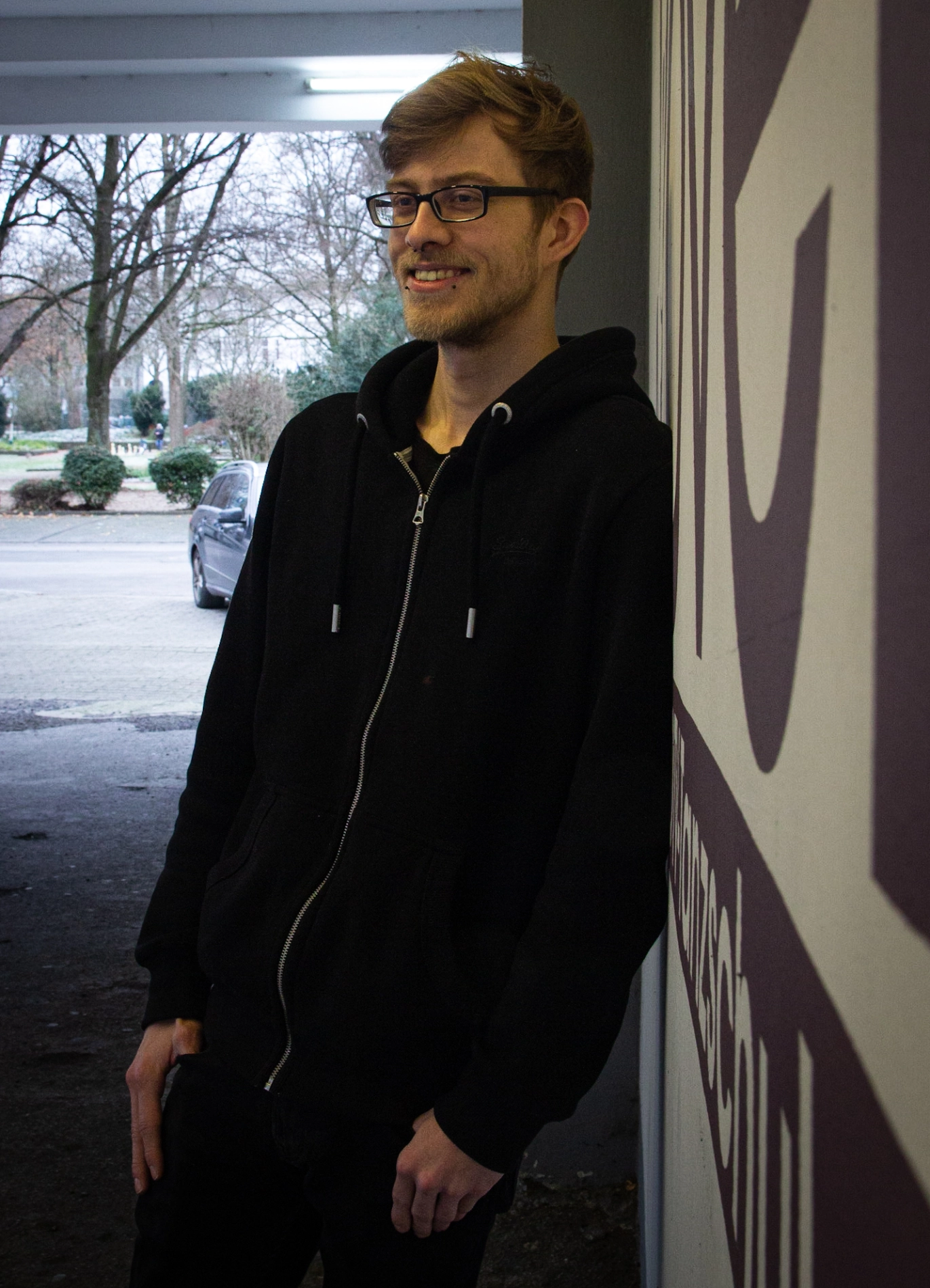 Lucas Schliepkorte Ein lächelnder Lucas Schliepkorte lehnt in schwarzem Hoodie mit Brille an einer Wand; draußen sind Straße, Bäume und ein geparktes Auto zu sehen.