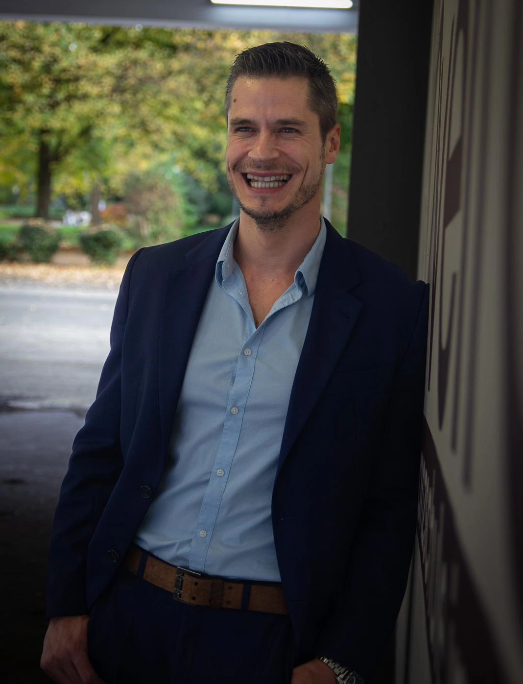 Sven-Dominique Bellinghausen Ein lächelnder Sven Dominique Bellinghausen steht im dunkelblauen Anzug mit hellblauem Hemd in einem hellen Innenraum vor grünem Hintergrund dort.